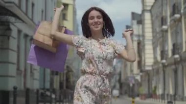 Alışveriş çantaları ile sevimli neşeli genç kadın. Şehir Sokağı boyunca dans ederken kulaklık ve cep telefonu ile kız müzik dinler. Açık havada zevk şık yaz elbisesi giyen genç eğlenceli kız.