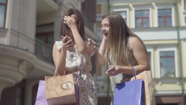 Zwei glückliche Freundinnen nach dem Einkauf mit Einkaufstüten, die auf dem Handy vor schönen Gebäuden SMS schreiben. Freizeitmädchen in stylischen Sommerkleidern genießen die gemeinsame Zeit. — Stockvideo