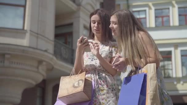 Due amiche carine dopo aver fatto shopping con le borse della spesa scattare selfie sul cellulare all'aperto. Tempo libero di ragazze felici. Signore spensierate a piedi attraverso la strada della città . — Video Stock