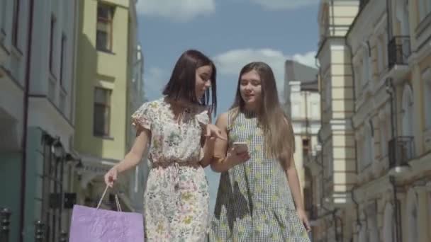 Due adorabili amiche dopo aver fatto shopping con le borse della spesa di fronte a splendidi edifici che discutono notizie guardando lo schermo del cellulare. Tempo libero di positive ragazze fashionista — Video Stock