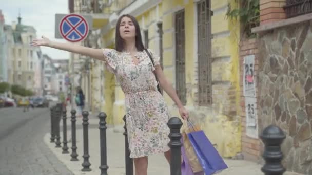 Mujer joven y despreocupada vistiendo vestido de verano con bolsas de compras en las manos tratando de coger el taxi en la calle de una vieja ciudad europea. Ocio de chica feliz. Tiempo de compras, vacaciones — Vídeos de Stock