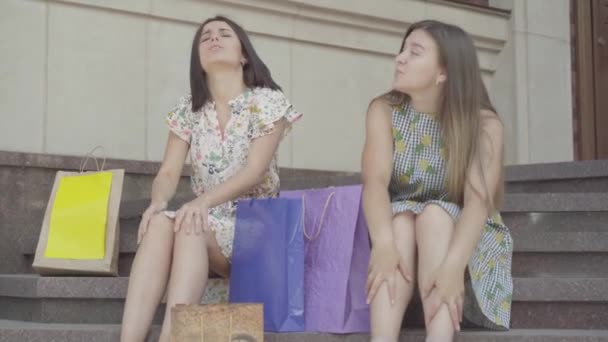 Dos novias atractivas sentadas en las escaleras después de comprar con bolsas de compras. Las damas están cansadas y agotadas. Ocio de chicas felices. Damas despreocupadas en la ciudad . — Vídeos de Stock