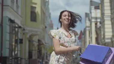 Alışveriş çantaları ile sevimli neşeli genç kadın. Şehir Sokağı boyunca dans ederken kulaklık ve cep telefonu ile kız müzik dinler. Açık havada zevk şık yaz elbisesi giyen genç eğlenceli kız.