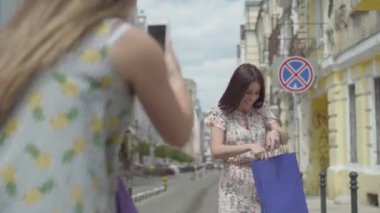 İki mutlu kız birlikte sokakta eğlenmek. Kız cep telefonunda arkadaşı fotoğraf çekerken dışarıda alışveriş yaptıktan sonra. Kız bir elbise üzerinde çalışıyor ve poz