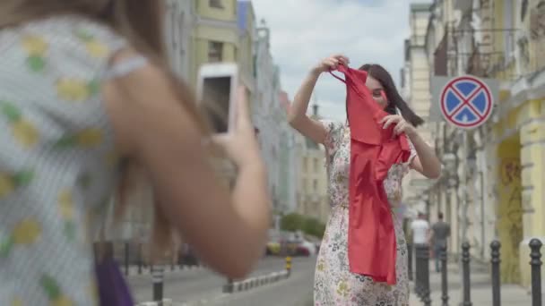 屋外でセル幸せな愛らしい楽しいファッショニスタガールフレンド上の女の子の写真。幸せな女の子は一緒に通りに立っている彼女の新しい赤いドレスを披露します。幸せな若い女性のレジャー. — ストック動画