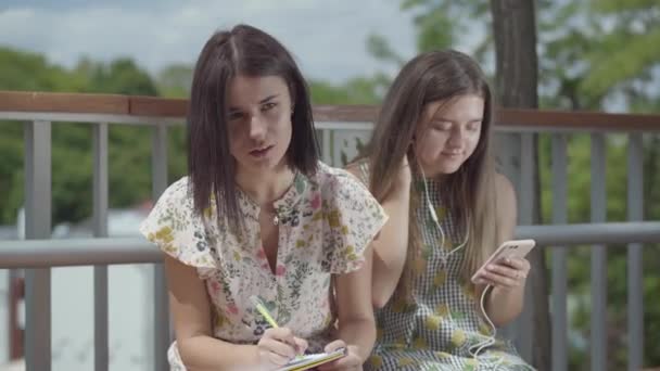 Két vonzó női hallgatók ülnek együtt a szabadban. Egy lány zenét hallgat vele fejhallgató, második egy írás le a jegyzetfüzetbe. Barátnői szórakozás. — Stock videók