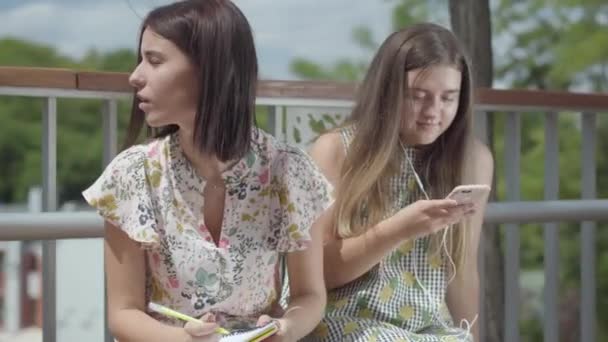 Dwie śliczne młode kobiety studenci siedzący na zewnątrz razem. Jedna dziewczyna słucha muzyki z jej słuchawek, drugi piśmie w swoim notebooku. Summertime wypoczynek koleżanek. — Wideo stockowe