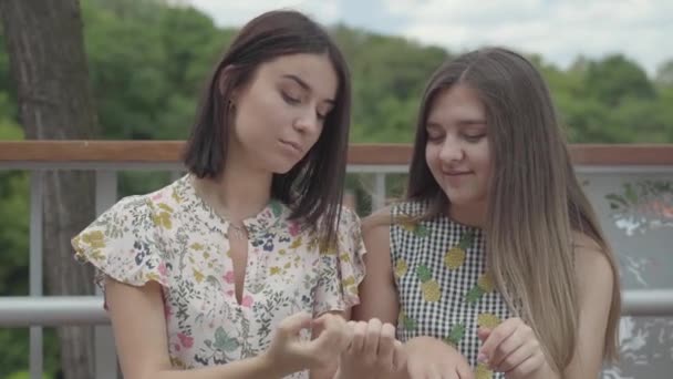 一緒に屋外に座っている2人の魅力的な女の子は、指と爪を示し、新しいマニキュアについておしゃべりしています。日常の事を話すガールフレンド。サマータイムレジャー — ストック動画