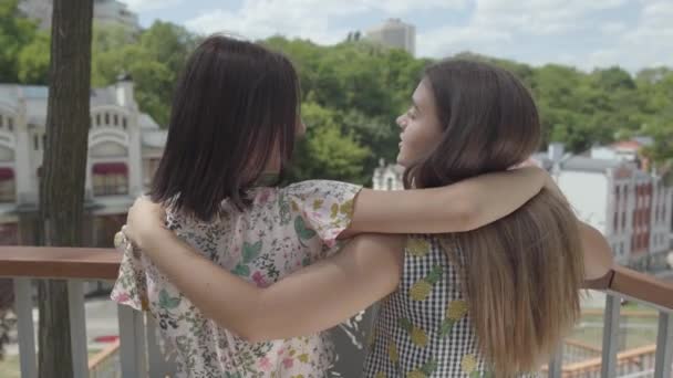 Dwie śliczne młode kobiety stojące na zewnątrz razem przytulanie i mówienie, podziwiając piękny pejzaż miejski. Dziewczyny na sobie Letnie sukienki mówią o codziennych sprawach. Summertime wypoczynek. Widok z tyłu — Wideo stockowe