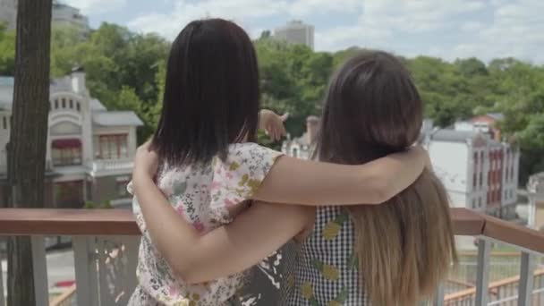 Två bedårande ung kvinna stående utomhus tillsammans kramas och pratar, beundra vackra stadsbilden. Flickvänner bär sommar klänningar talar om vardagliga frågor. Sommartid fritid. Back View — Stockvideo