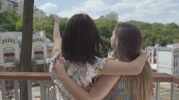 Deux jolies filles debout à l'extérieur ensemble étreignant et parlant, admirant le beau paysage urbain. Des copines discutant d'architecture. Loisirs d'été. Vue arrière — Video