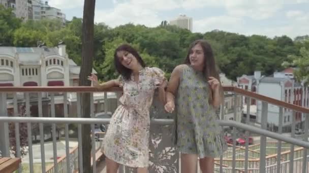 Dos chicas atractivas bailando al aire libre juntas frente a un hermoso paisaje urbano. Novia despreocupada divirtiéndose al aire libre. Tiempo libre de verano — Vídeos de Stock