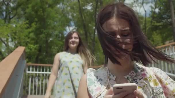 Atrakcyjna cute młoda dziewczyna siedzi na pierwszym planie tekstylny na telefon komórkowy. Beztroki przyjaciel biegnie za nią i zamyka jej oczy przyjaciół z rękami, koleżanki przytulanie. Koncepcja przyjaźni — Wideo stockowe