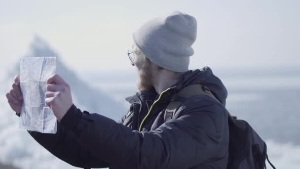 Портрет молодого белокурого бородатого красавца в теплой куртке и шляпе, стоящего на леднике, сверяясь с картой. Удивительная природа снежного Северного или Южного полюса. Турист перед льдом — стоковое видео