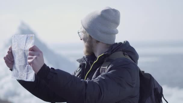 Retrato de jovem loiro barbudo bonito vestindo casaco quente e chapéu em pé na geleira verificando com o mapa. Natureza surpreendente de um Norte nevado ou Pólo Sul. O turista em frente ao gelo — Vídeo de Stock