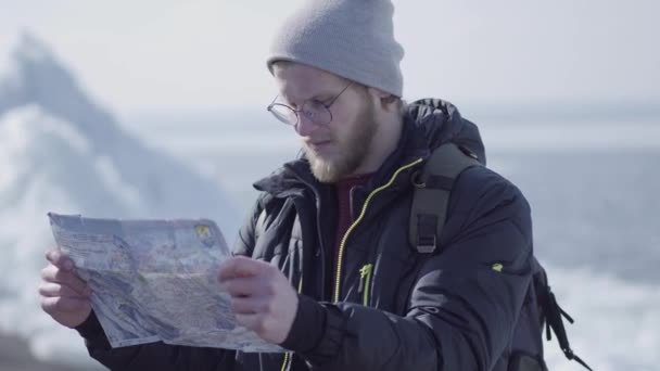 Retrato de un joven rubio barbudo hombre guapo con chaqueta caliente y sombrero de pie en el glaciar comprobando con el mapa. Increíble naturaleza de un nevado Polo Norte o Sur. El turista frente al hielo — Vídeo de stock