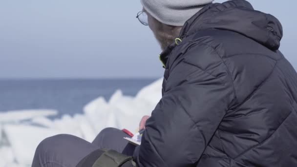 Un joven con una chaqueta cálida y un sombrero sentado en el glaciar escribiendo sus pensamientos en un pequeño cuaderno. Increíble naturaleza de un glaciar nevado. El explorador polar en el bloque de hielo — Vídeos de Stock