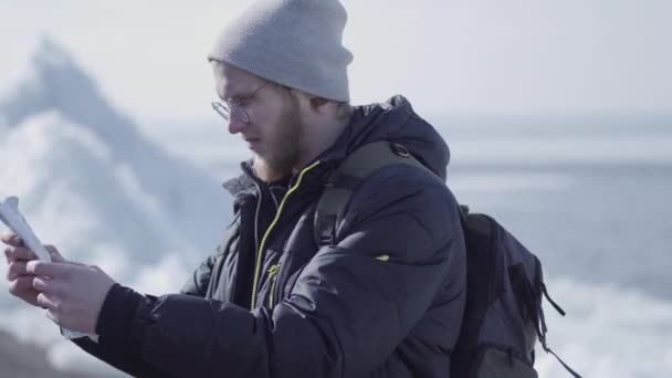 Handsome blond bearded man in glasses with backpack checking the map in front of ice at the North or South pole. Tourist travels in the winter — Stock Video