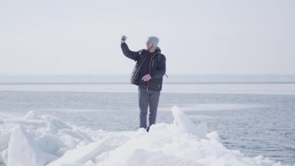 Knappe blonde bebaarde man in glazen in warme winterjas staande op het ijsblok bij gletsjer het nemen van foto op mobiele telefoon. Tourist Travels concept — Stockvideo