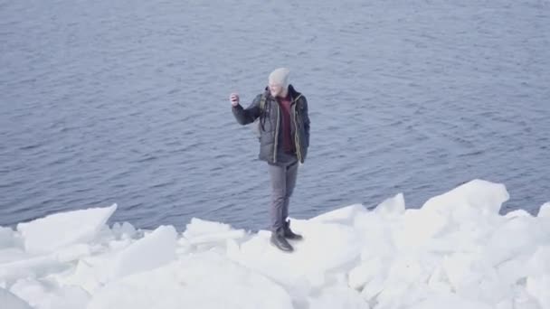 Hombre guapo con gafas en abrigo de invierno caliente de pie en el bloque de hielo en el glaciar tomando fotos en el teléfono celular. Concepto de viajes turísticos — Vídeo de stock