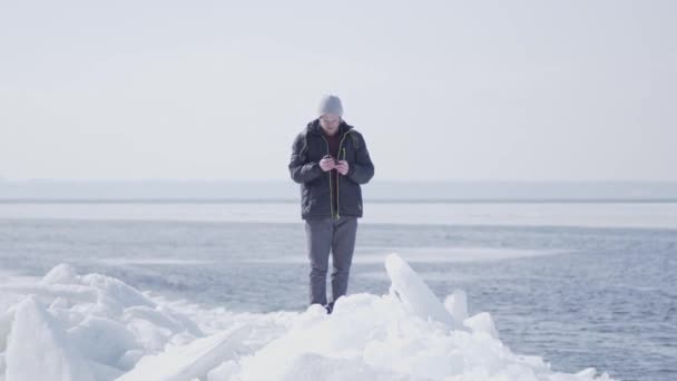 Hombre guapo y rubio barbudo con gafas de abrigo de invierno parado en el bloque de hielo en el glaciar tomando fotos en el teléfono celular. Viajes turísticos en invierno — Vídeos de Stock
