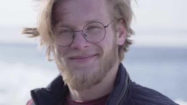 Portréja csinos szakállas férfi szemüveg ragadt szőke haja keres a kamera mosolygós közelről. Pozitív fickó, elöl kilátás — Stock videók