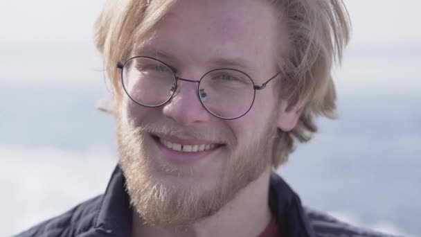 Retrato de primer plano del guapo barbudo con gafas de pelo rubio mirando a la cámara sonriendo al aire libre. Ocio chico positivo . — Vídeo de stock