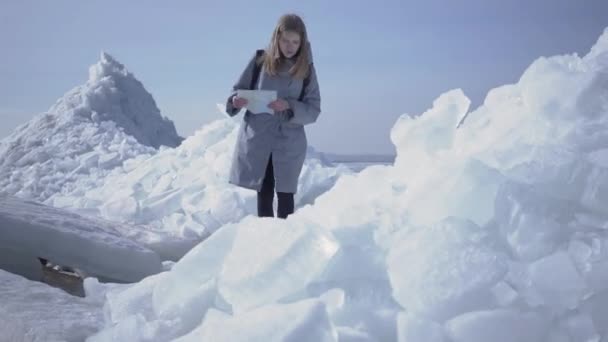 Joven mujer bonita rubia triste con chaqueta caliente deslizándose mientras camina sobre el glaciar con mapa en las manos. Increíble naturaleza de nieve Polo Norte o Sur. El turista rodeado de bloques de hielo. La señora está perdida — Vídeo de stock