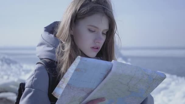 Retrato de una joven rubia guapa con una chaqueta cálida parada sobre el glaciar con el mapa en las manos, mirando a su alrededor. El turista frente a los bloques de hielo. La señora está perdida y tratando de encontrar una manera — Vídeo de stock