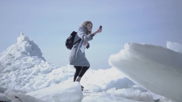 Νεαρή ξανθιά όμορφη γυναίκα με ζεστό μπουφάν περπατάει στον παγετώνα με το χάρτη στα χέρια, κρατώντας το κινητό της σε ανυψετό χέρι, προσπαθώντας να βρει το δίκτυο κινητής τηλεφωνίας. Ο τουρίστας μπροστά από τα τετράγωνα πάγου — Αρχείο Βίντεο