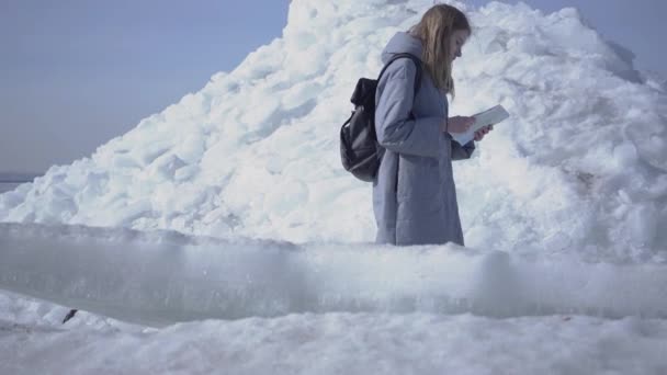 Młoda smutna blond ładna kobieta w ciepłym płaszczu poślizgnięcia podczas chodzenia na lodowcu z mapą w rękach. Niesamowita natura śnieżnych bieguna północnego lub południowego. Turysta otoczony przez bloki lodu. Lady jest stracone — Wideo stockowe