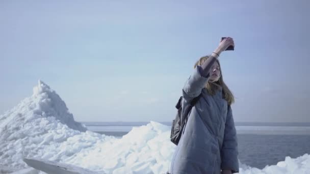 Schattige jonge vrouw in warme jas wandelen op de gletsjer, houden haar mobiele telefoon in verhoogde hand, proberen om mobiel netwerk te vinden. De toerist voor de ijsblokken en de zee — Stockvideo