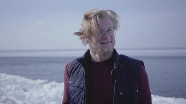 Zbliżenie portret atrakcyjny młody blond człowiek w okularach patrząc od dotykając jego włosy. Atrakcyjny facet skandynawskiego wyglądu przed zimowym krajobrazem na biegunie północnym lub południowym — Wideo stockowe