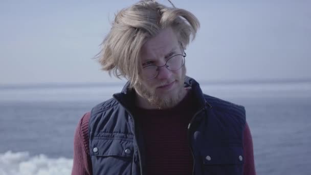 Gros plan portrait de beau jeune homme blond dans des lunettes regardant ailleurs toucher ses cheveux. gars attrayant de l'apparence scandinave en face du paysage hivernal au pôle Nord ou Sud — Video