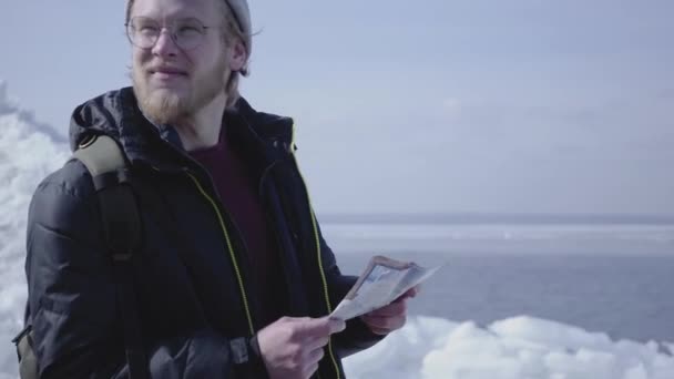 Retrato del joven rubio barbudo guapo vestido con chaqueta caliente y sombrero caminando por el glaciar comprobando con el mapa. Increíble naturaleza de un nevado Polo Norte o Sur. El turista frente al hielo — Vídeo de stock