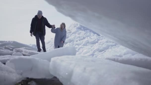 Knappe bebaarde man lopen met mooie vrouw handen vasthouden, de man helpt zijn vriendin om de ijsblokjes te beklimmen.. Prachtig uitzicht op een besneeuwde Noord-of Zuidpool. Een paar toeristen op de gletsjer — Stockvideo