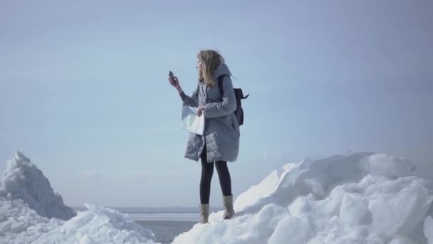 Atrakcyjna blond kobieta z mapą próbującą złapać sieć posiadając telefon z wysokim stojąc na bloku lodu na biegunie północnym lub południowym. Dziewczyna jest zagubiona i chce znaleźć sposób — Wideo stockowe