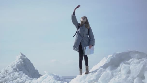 Portrait of cute woman in warm jacket walking on the glacier, holding her cellphone in raised hand, trying to find mobile network. The tourist in front of the ice blocks and sea — Stock Video