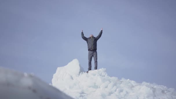 Emocional hombre feliz con abrigo caliente de pie en la parte superior del glaciar vomitando sus manos, triunfando. Increíble vista de un nevado Polo Norte o Sur. El macho está en el bloque de hielo. El tipo llegó a la cima. — Vídeo de stock