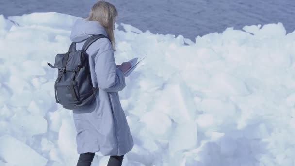 Młoda blond ładna kobieta ubrana w ciepłą kurtkę stojącą na lodowcu sprawdzając mapę. Niesamowita natura śnieżnych bieguna północnego lub południowego. Turysta przed bloków lodu. Zwolnionym. — Wideo stockowe