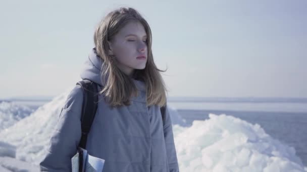 Portrait de jeune jolie femme blonde vêtue d'une veste chaude debout sur le glacier avec la carte dans les mains, regardant autour. Le touriste devant les blocs de glace. Mouvement lent . — Video