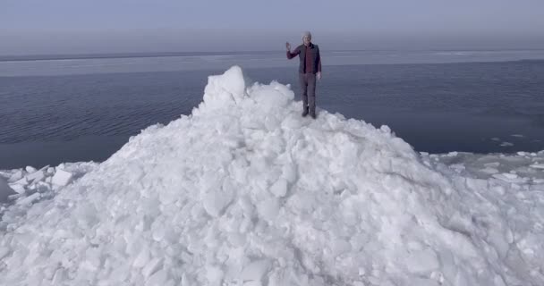 Вид на повітряний дрон молодого активного щасливого чоловіка, який перебуває на льодовикових льодовиках біля берегової лінії зимового моря, тремтячи рукою. Перегляд безпілотника — стокове відео