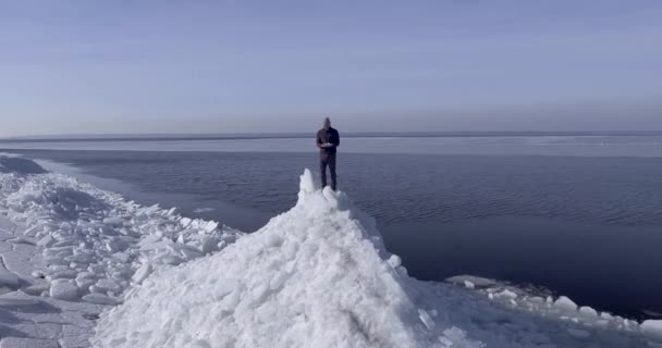 Vista aérea del dron del joven hombre feliz activo que permanece en los glaciares de hielo cerca de la costa del mar de invierno. Drone vuela en las manos de un chico . — Vídeo de stock