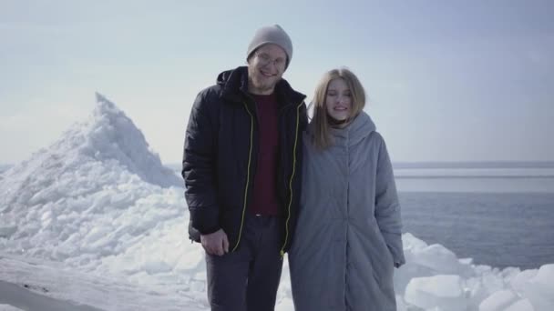 英俊的金发男子和迷人的年轻女子在温暖的外套手牵手。背景上雪白的北极或南极的迷人景色。一对游客正在探索冰川。微笑的人 — 图库视频影像