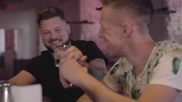 Due uomini adulti battono i bicchieri al bancone del bar sorridendo e parlando. Amici maschi rilassati che si rilassano insieme bevendo alcol d'elite. Giorno libero — Video Stock
