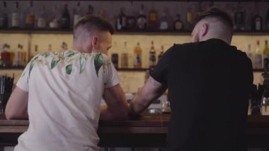 Barın tezgâhında oturan iki adam elit alkol içiyor. Gün izin.