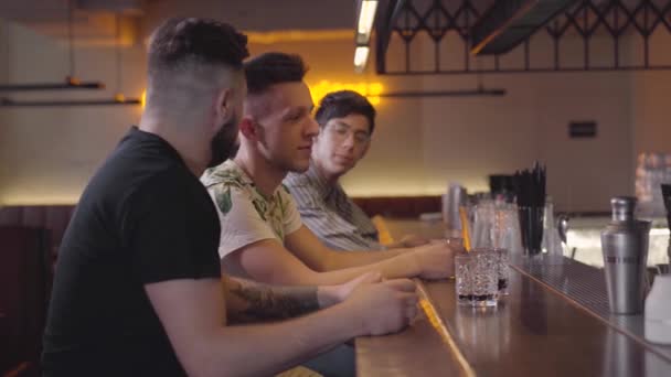 バーカウンターに座っている2人の大人の男性、会社に入社した3人目の男は、仲間が抱き合い、ハイ5を与える。リラックスした男性の友人は、一緒にエリートアルコールを飲んで冷やします。休み日 — ストック動画