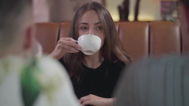 Linda chica sentada frente a dos amigos varones tomando café. Mujer joven y dos chicos se comunican sentados en una mesa en un restaurante . — Vídeos de Stock