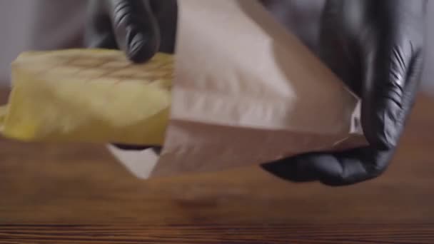 El cocinero en guantes de cocina negros pone el rollo de riego en una bolsa de papel. Concepto de publicidad alimentaria. Delicioso sándwich o shawarma listo para comer — Vídeo de stock