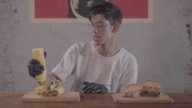 Sakallı adam iştah açıcı burger ve shawarma yemek için hazırlanıyor önünde masada oturuyor. Adam sandviçin üzerine lezzetli sos döküyor..